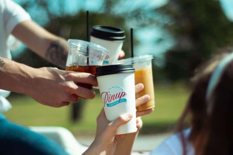 Four people clinking coffee cups outdoors, holding a mix of iced and hot beverages. Trees and grass are visible in the background.