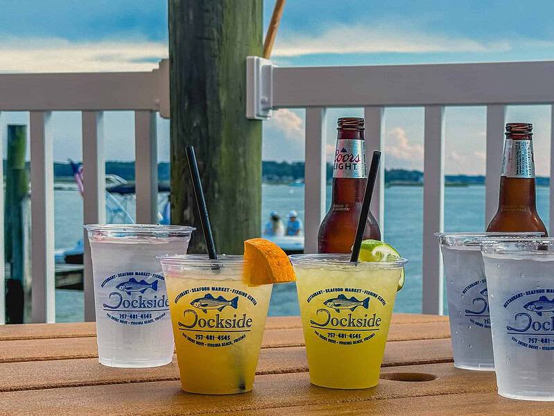 Drinks on a wooden table by the water, including two beers, bottled water, a citrus cocktail, and a cucumber drink. A person holds a phone in the background. Dockside logo on cups.