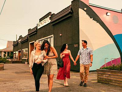 Two women walk arm in arm, followed by another woman and a man holding hands, outside a building with a mural.