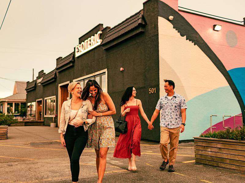 Two women walk arm in arm, followed by another woman and a man holding hands, outside a building with a mural.