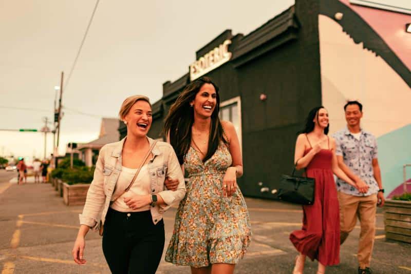 Two women walk and laugh in the foreground, with a couple walking behind them. They are outdoors near a building with wall art.