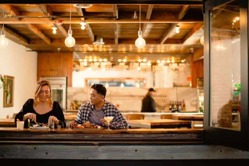 Two people sit at a bar-style counter in a warmly lit restaurant, eating and drinking. Hanging bulbs illuminate the wooden interior, and another person is visible in the background.