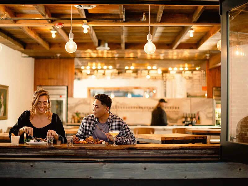 Two people sit at a wooden counter in a restaurant, eating and chatting. Hanging lights illuminate the scene. A person works in the background.