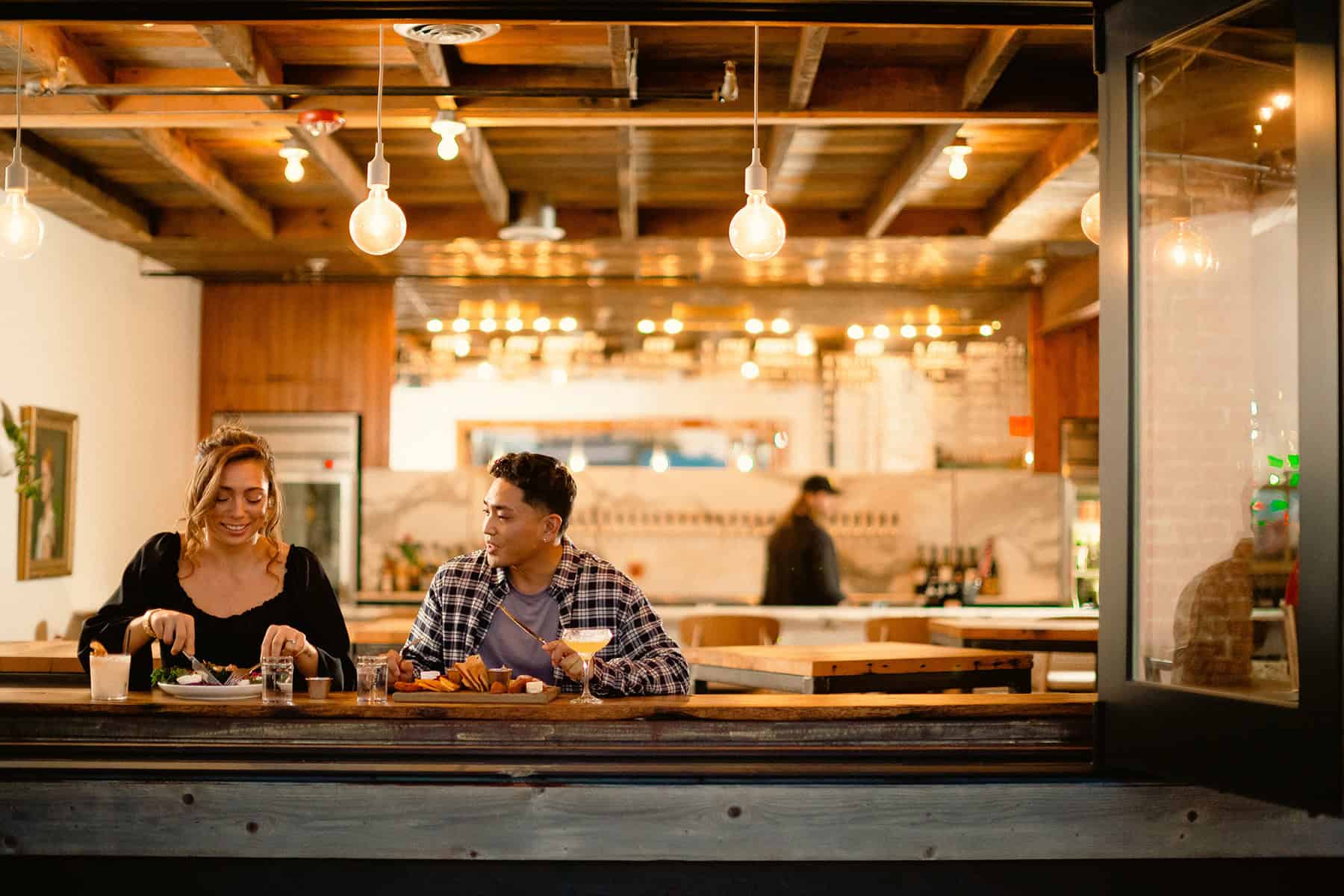 Two people sit at a wooden counter in a restaurant, eating and chatting. Hanging lights illuminate the scene. A person works in the background.
