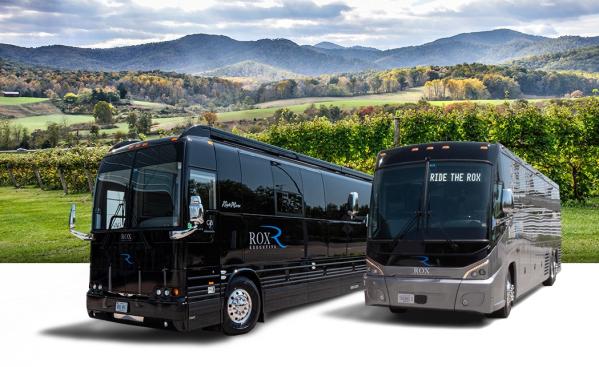 Two black tour buses labeled "ROX" parked side by side in front of a scenic landscape with rolling hills and lush greenery.