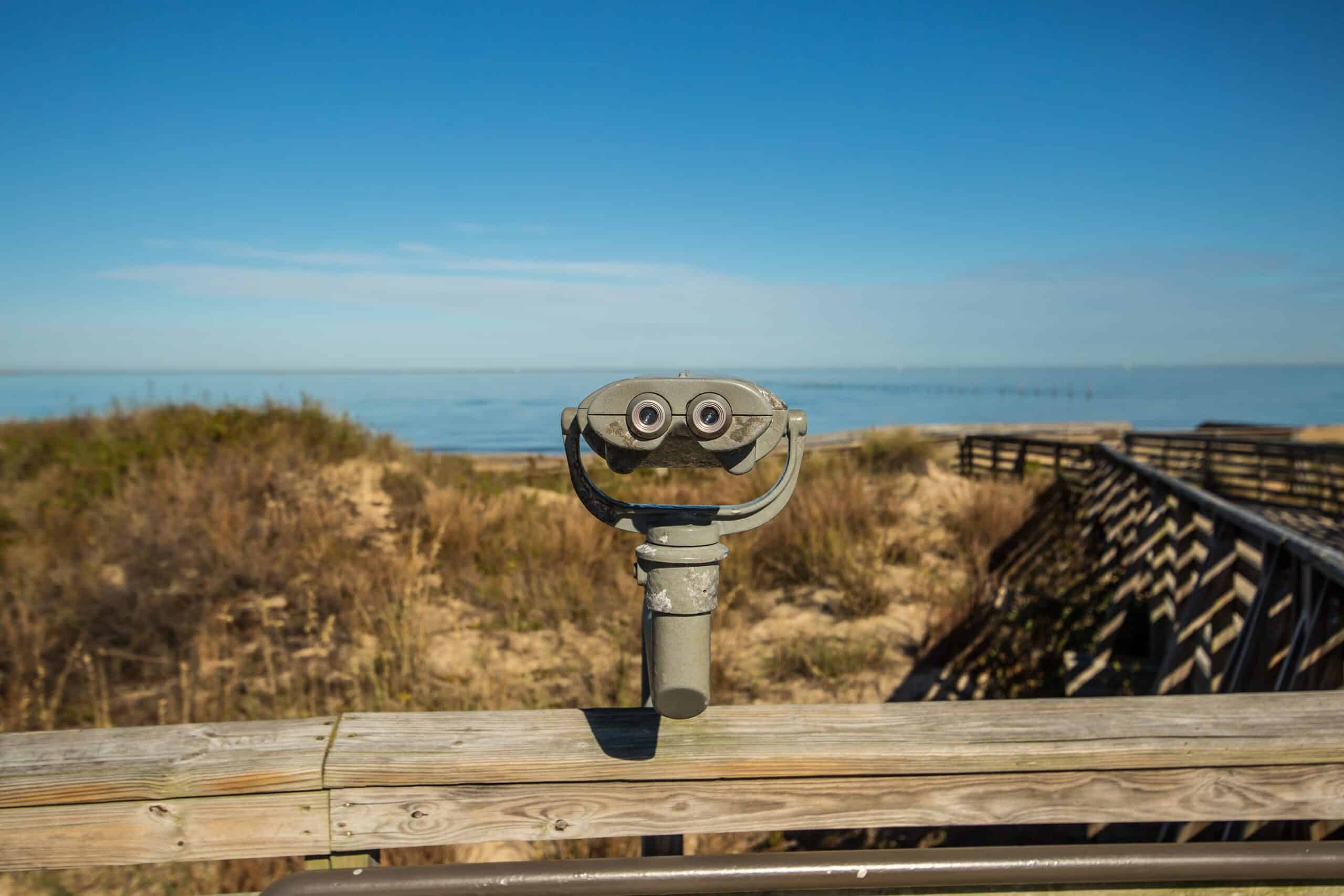 Mounted binoculars on a wooden railing overlook Virginia Beach with sandy dunes and a clear blue sky.