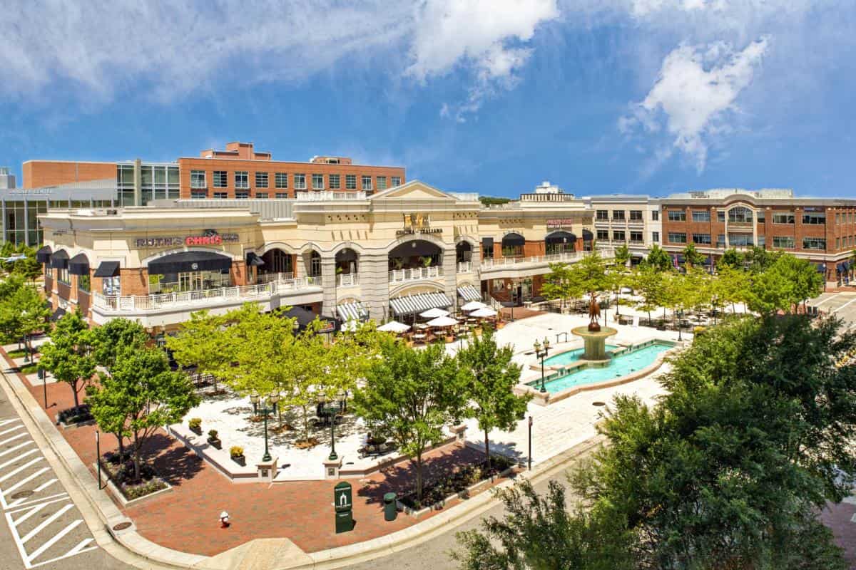 Outdoor shopping center with trees, a fountain, and people walking. Buildings house various stores and restaurants under a blue sky.
