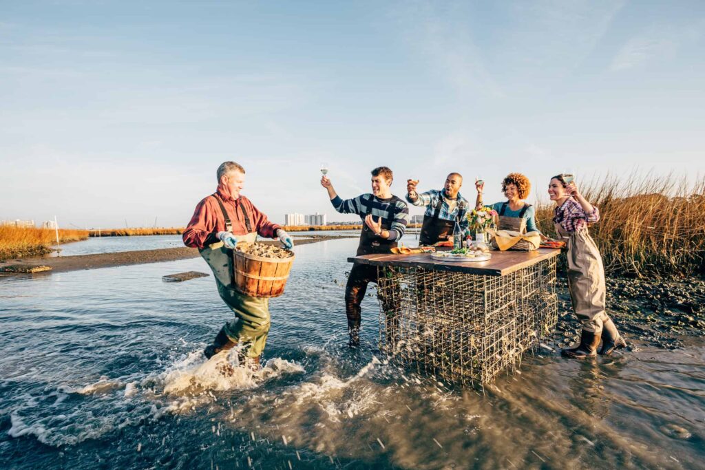 A group of people in waders stand around a table in shallow water at the Pleasure House Oyster Farm. One person is carrying a basket of fresh oysters. They appear to be toasting with glasses.