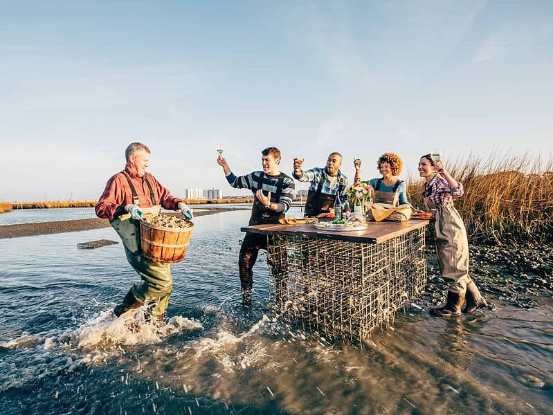 A group of people in waders stand around a table in shallow water. One person is carrying a basket. They appear to be toasting with glasses.