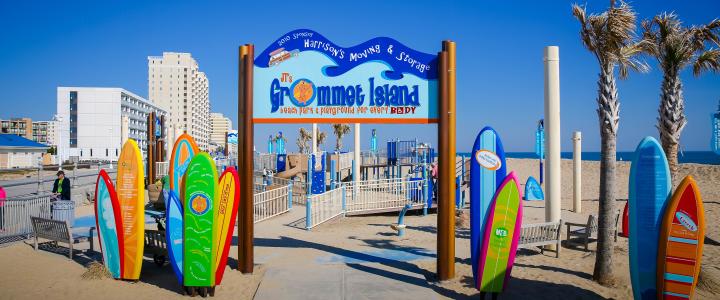 Entrance to Grommet Island park with surfboard-shaped signs and a blue sky. Buildings and palm trees are in the background.