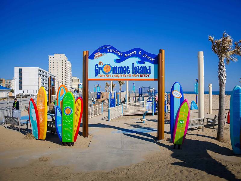 Entrance gate to Grommet Island Park, a beach playground in Virginia Beach, with colorful surfboards and palm trees under a clear blue sky.