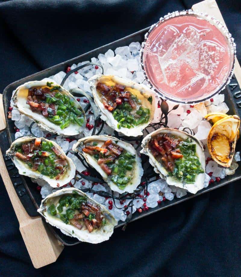 Tray of oysters topped with greens and bacon on ice, served with a pink cocktail and grilled lemon wedges.