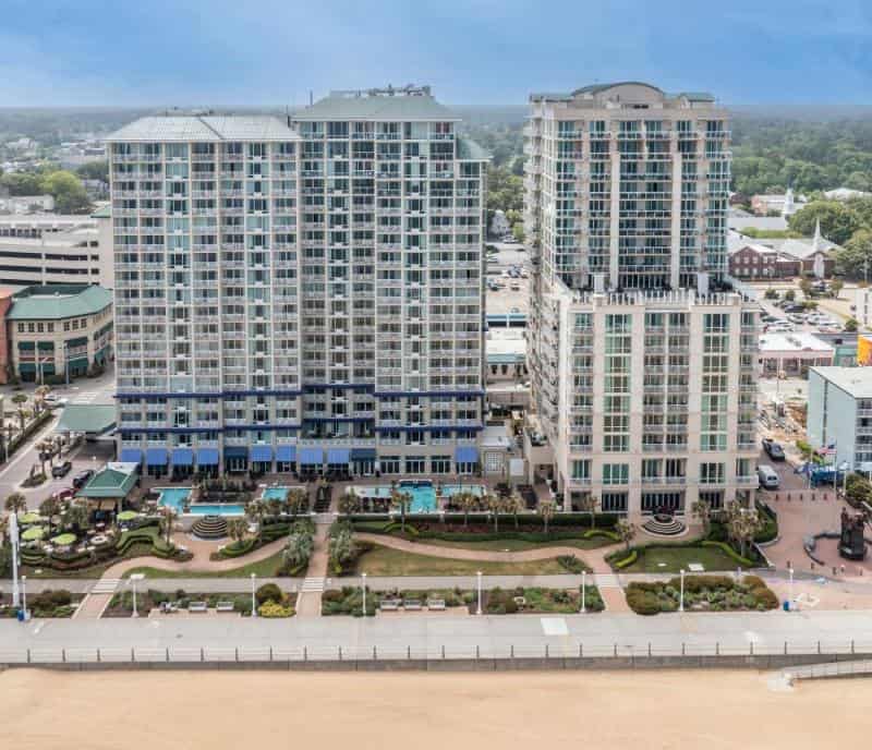 Two tall, modern beachfront buildings with balconies, situated near a landscaped area and boardwalk, overlooking a sandy beach and ocean. The town and greenery are visible in the background.