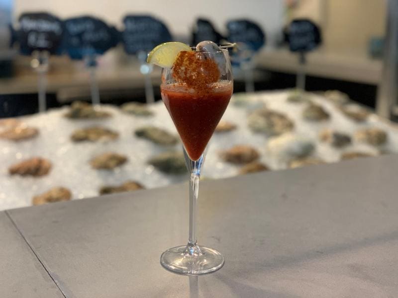 A red cocktail garnished with a lime wedge and olives sits on a counter, with blurred signage and items in the background.