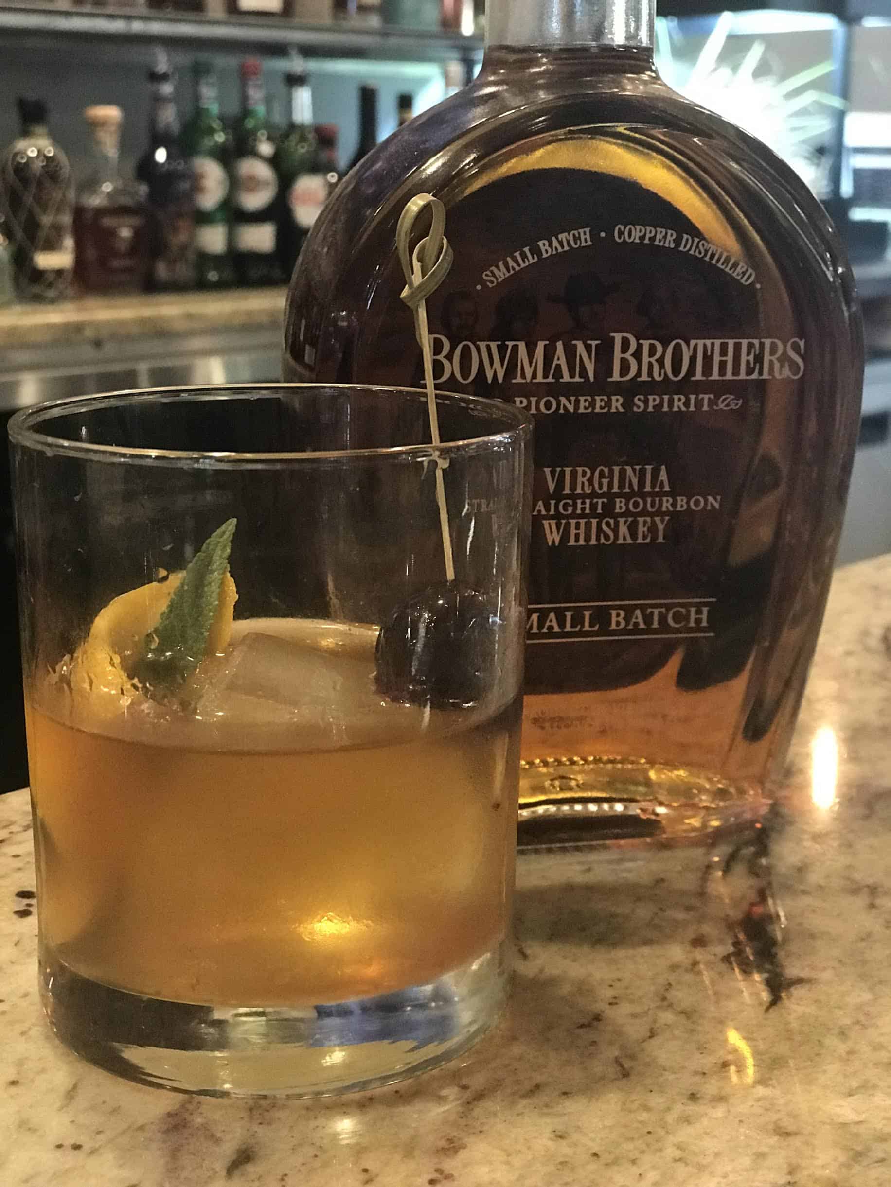 A glass of Old Fashioned cocktail with a cherry and lemon twist garnish sits beside a bottle of Bowman Brothers Virginia Straight Bourbon Whiskey on a marble counter.