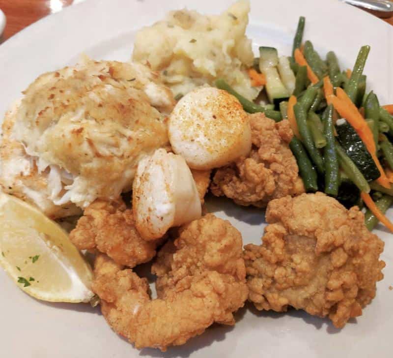 A plate with a crab cake, fried oysters, scallop, mashed potatoes, and mixed vegetables including green beans and carrots. Garnished with a lemon wedge.