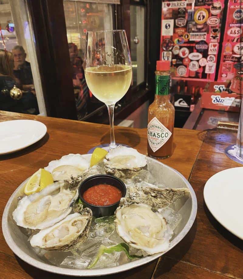 A plate with six oysters on ice, lemon wedges, and cocktail sauce. A bottle of Tabasco and a glass of white wine are beside it on a wooden table.