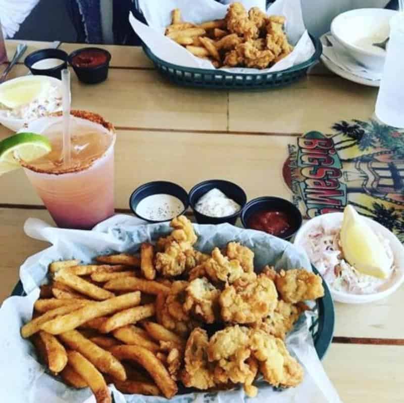Fried seafood with fries and coleslaw in baskets, four dipping sauces, and a pink cocktail on a wooden table.