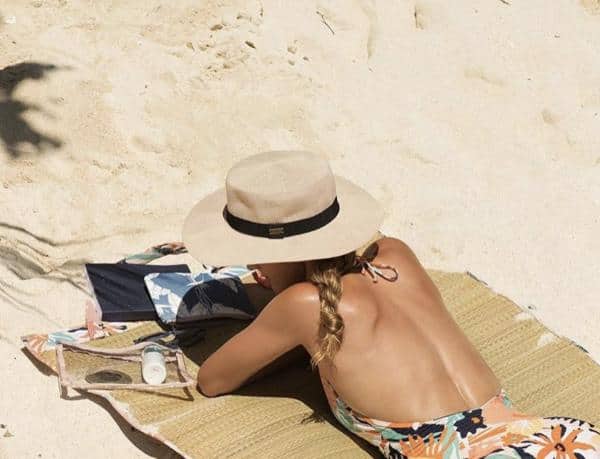 Person lying face down on a beach towel in the sand, wearing a sun hat and a floral swimsuit. A clear bag with sunscreen and sunglasses is nearby.