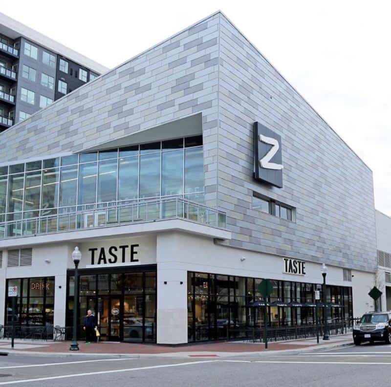 Modern building with "Taste" restaurant on the ground floor, large "Z" sign on the upper section, glass windows, and street lamps in front.