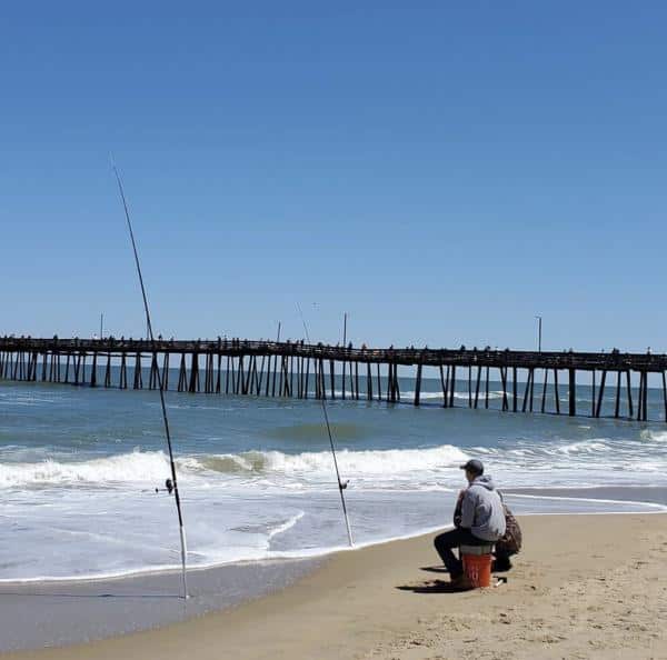 Person sitting on a bucket fishing on a sandy beach with two fishing rods in the sand. A pier extends into the water under a clear blue sky.