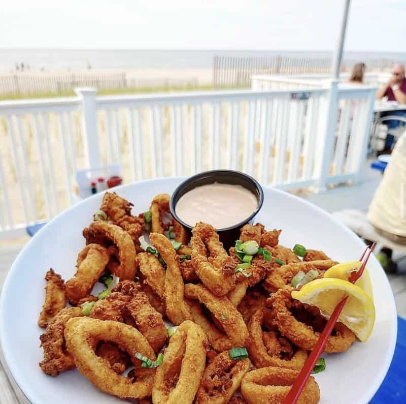 A plate of fried calamari with green onions and lemon wedges, served with dipping sauce, on an outdoor table overlooking a beach.