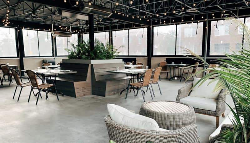 Modern outdoor patio with tables, chairs, and sofas. Overhead string lights create a cozy atmosphere. Potted plants add greenery.