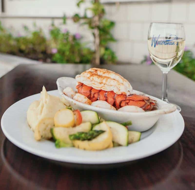 A plate with lobster tail, mashed potatoes, and mixed vegetables next to a glass of white wine on a table.