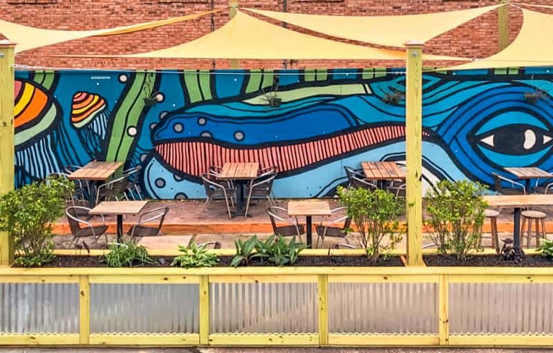 Outdoor seating area with wooden tables and metal chairs in front of a colorful mural featuring abstract shapes and designs.