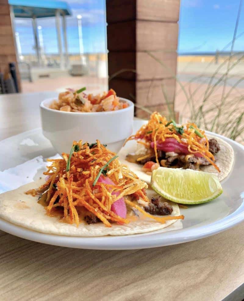 Two tacos topped with shredded vegetables on a plate, accompanied by a lime wedge and a bowl of slaw. Background shows an outdoor patio with beach and sky.