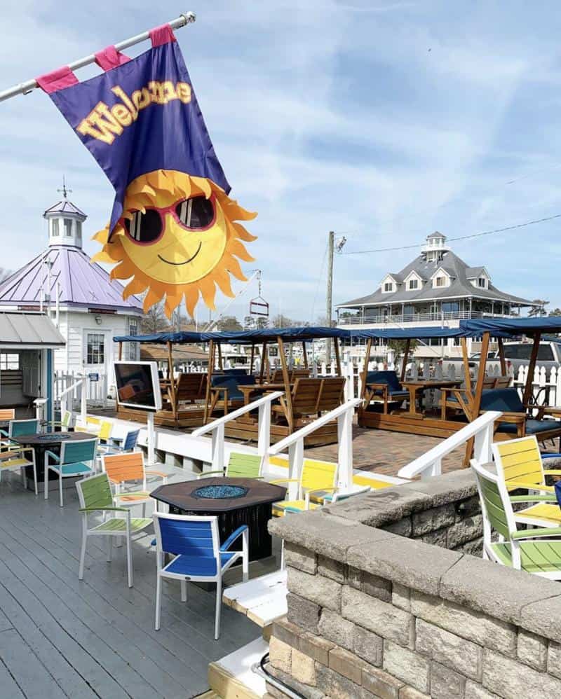 Outdoor seating area with colorful chairs and tables, featuring a "Welcome" flag with a smiling sun design. A lighthouse-style building is in the background.