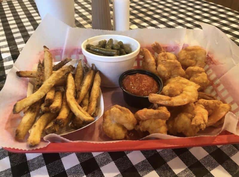 A basket with fried shrimp, French fries, and a small cup of green beans on a checkered tablecloth. Ketchup is served in a small cup.