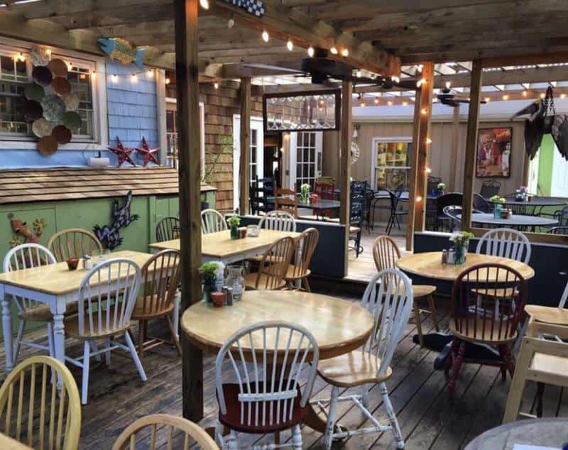Outdoor patio with wooden tables and mixed chairs under a pergola. String lights hang above, and decorative wall art is visible.