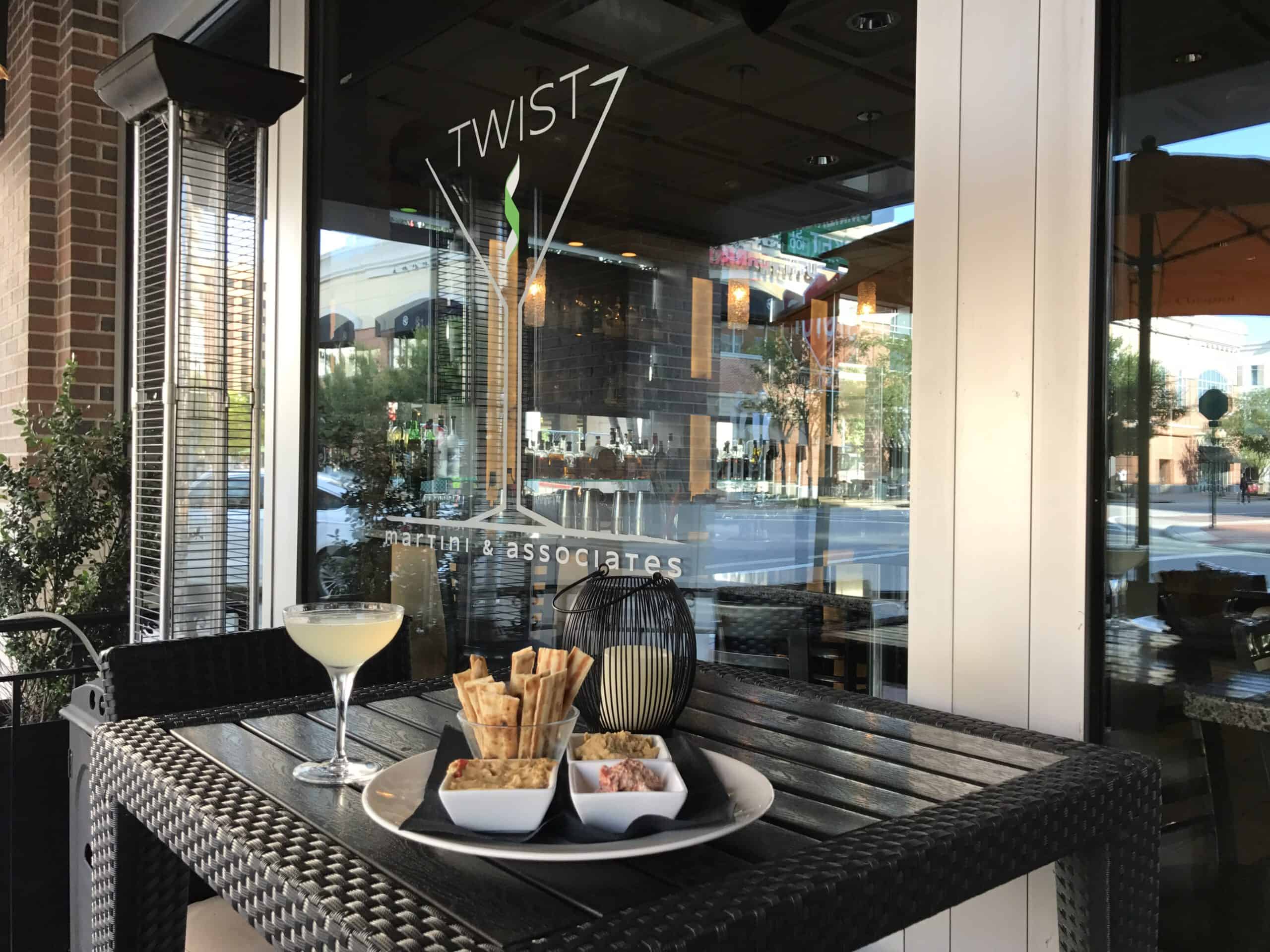 Outdoor table with a cocktail, crackers, and spreadable dip in front of a restaurant window with "TWIST" signage.