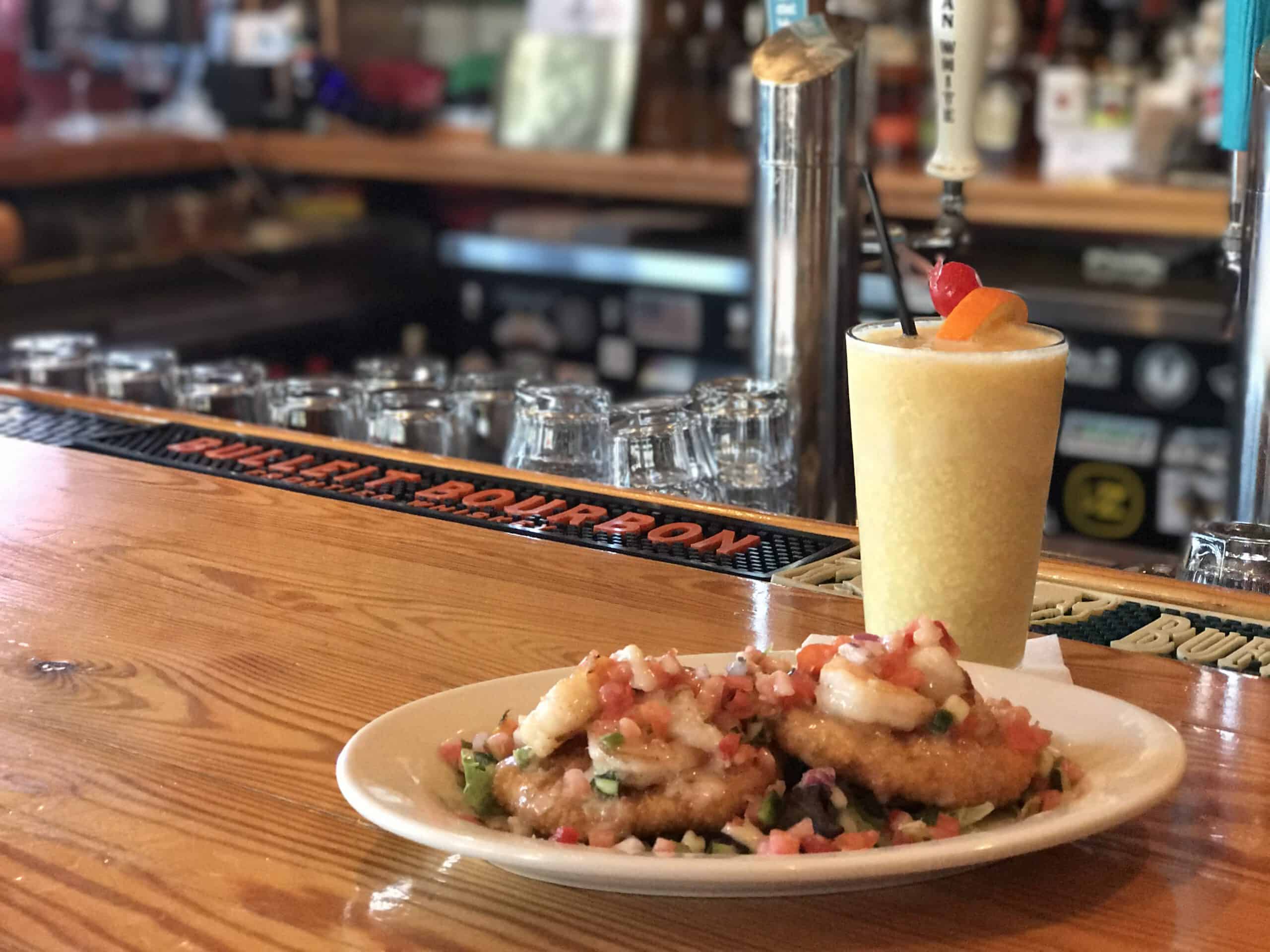 A plate of shrimp-topped appetizers on a wooden bar counter next to a frosty, garnished tropical drink.