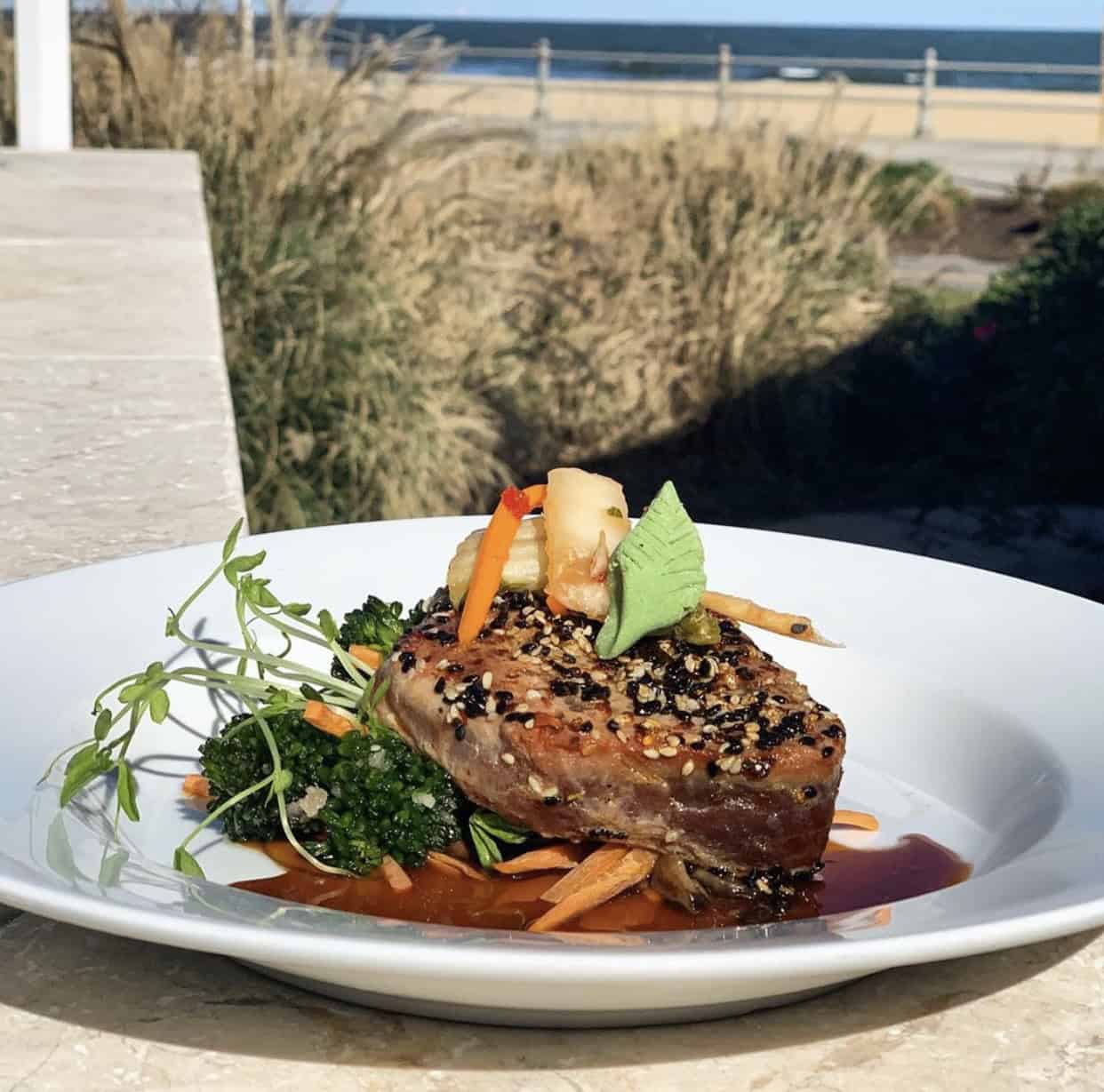 Plate of sesame-crusted tuna with greens, carrots, and sauce, garnished with herbs and apple slices. Beach in the background.