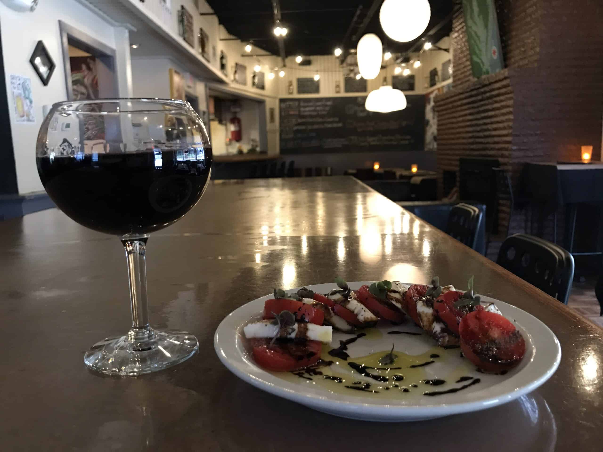 A glass of red wine next to a plate of caprese salad with tomatoes, mozzarella, and balsamic drizzle on a table in a dimly lit restaurant.
