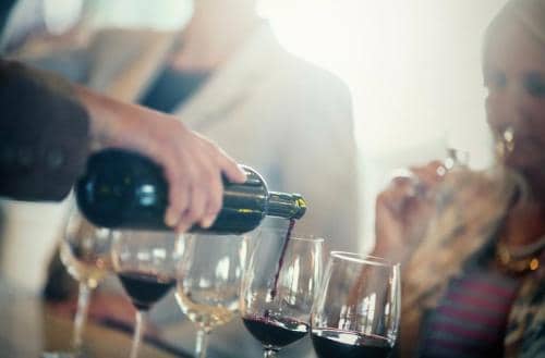 A person pours red wine into glasses while another person tastes wine.