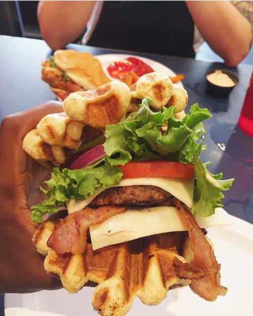 Waffle sandwich with lettuce, tomato, cheese, bacon, and burger patty. Person holding the sandwich at a table with a plate and a drink nearby.