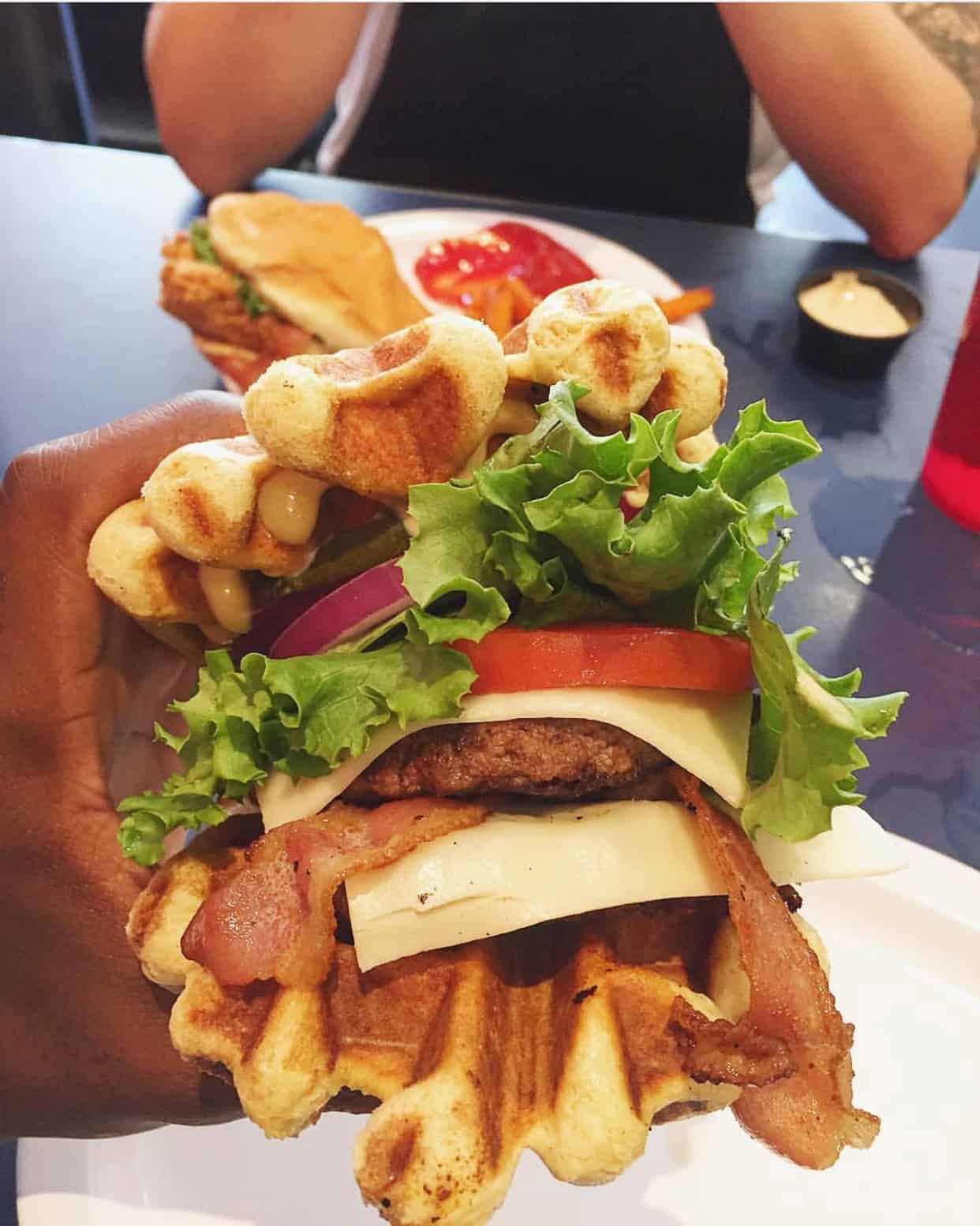 A waffle burger with lettuce, tomato, onion, cheese, bacon, and a beef patty, served with a side of sauce and a sandwich in the background.