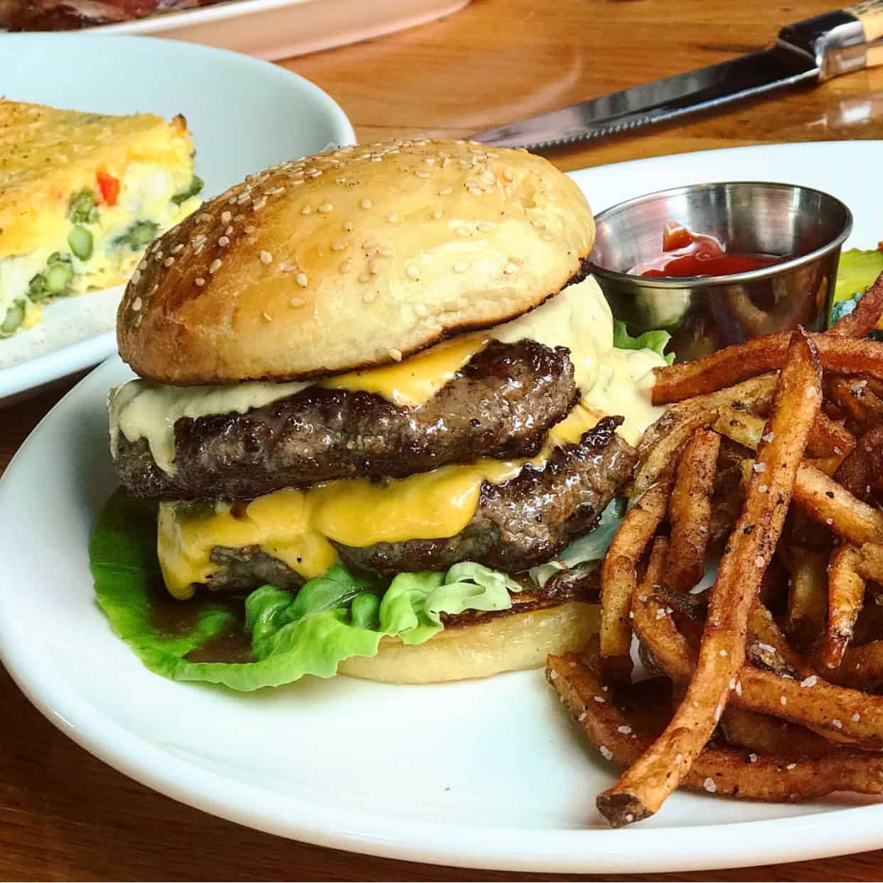 A cheeseburger with two beef patties, lettuce, and sauce, served with a side of fries and a cup of ketchup on a white plate.