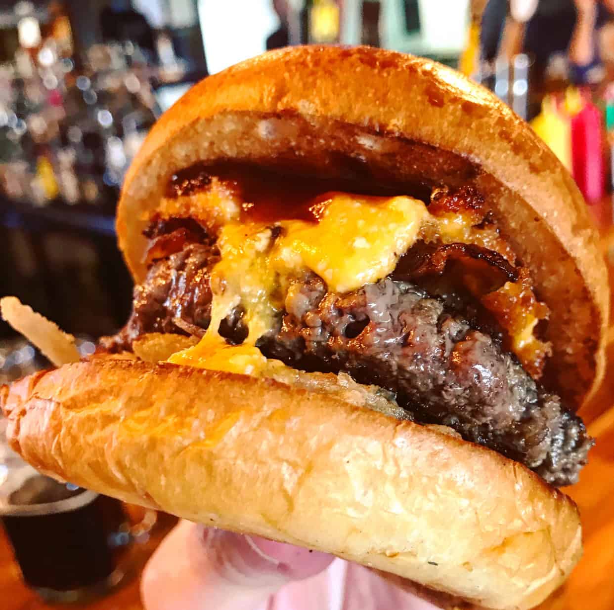 Close-up of a cheeseburger with melted cheddar, beef patty, bacon, and sauce in a toasted bun. Background shows a blurred bar setting.