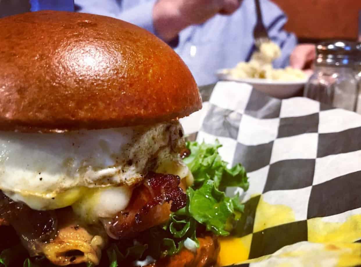 Close-up of a gourmet burger with a fried egg, bacon, lettuce, and cheese on a bun, set on a checkered paper. A person in the background is eating from a bowl.