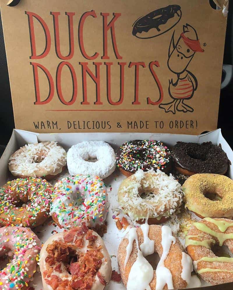A box of assorted Duck Donuts featuring various toppings like sprinkles, powdered sugar, bacon, and coconut under a branded lid.