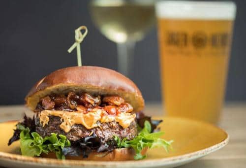 Burger with lettuce, cheese, and sauce on a plate, secured with a skewer. A glass of beer and a glass of wine are in the background.