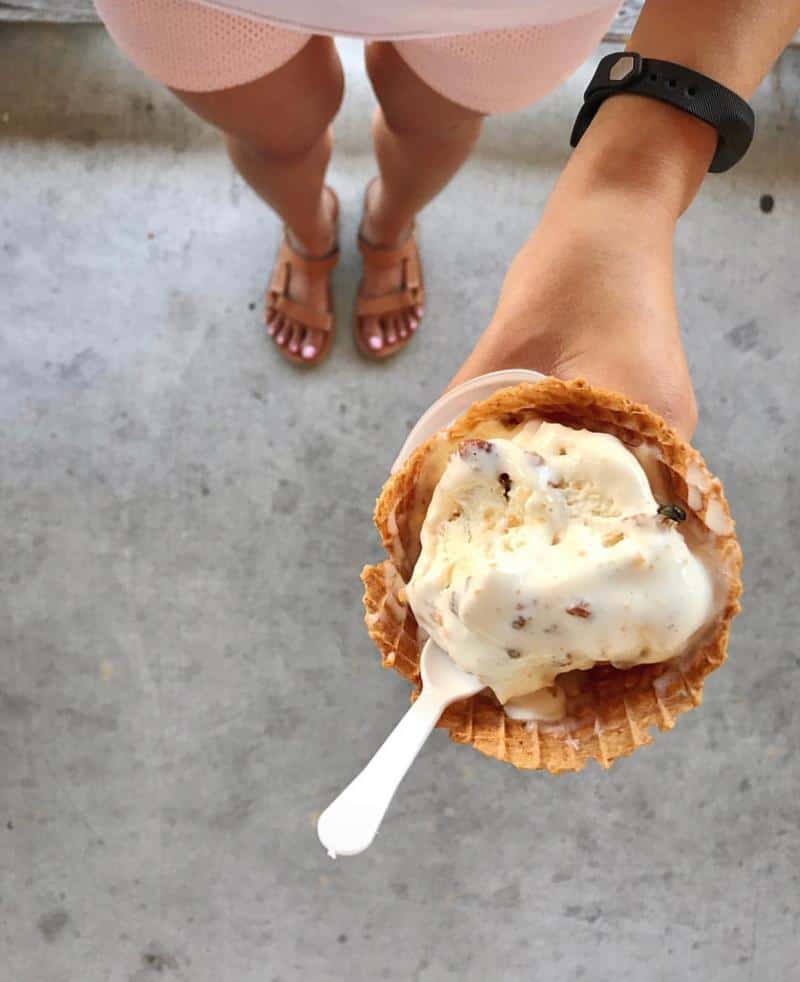 Person holding a waffle cone with vanilla ice cream seen from above, wearing a wristband and sandals.