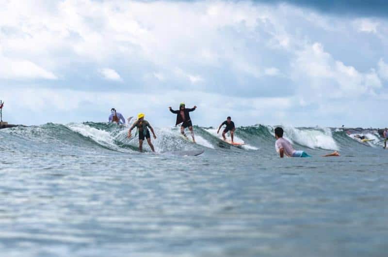 Several people are surfing on small waves under a cloudy sky. One person is lying on their board in the water.