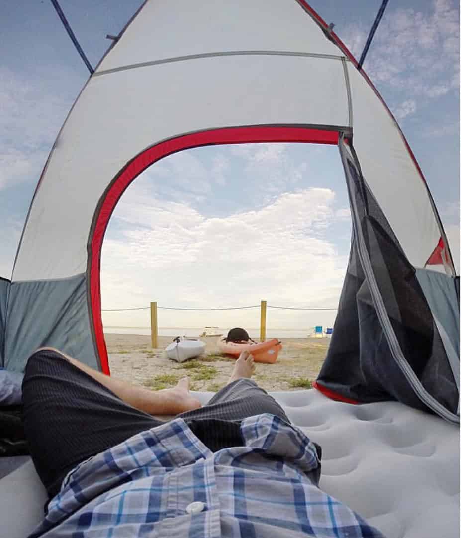 View from inside a tent facing the beach. A person lies on an inflatable mattress, wearing shorts and a plaid shirt. The ocean and a cloudy sky are visible through the tent's open flap.