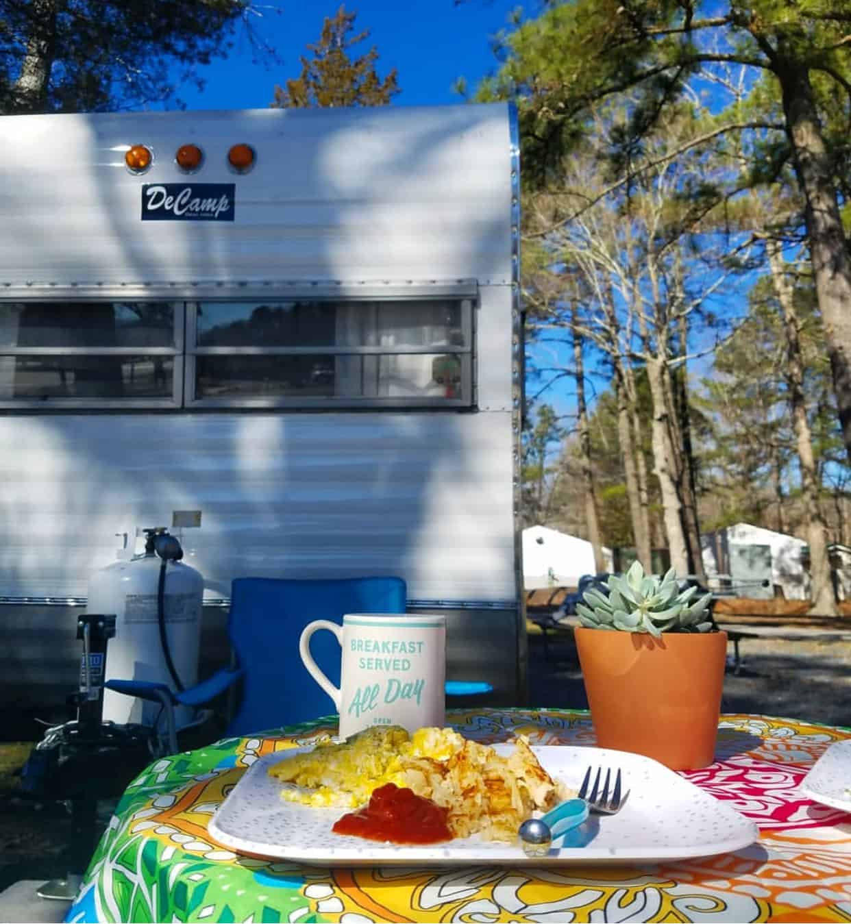Plate of eggs and hash browns with ketchup, a mug, and a potted plant on a colorful tablecloth outside a camper in a wooded campsite.