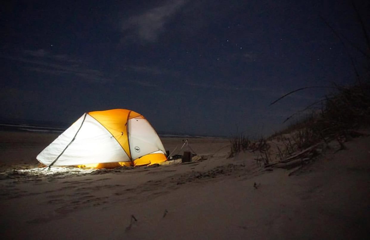 A glowing orange and white tent is set up on a sandy beach at night under a starry sky.